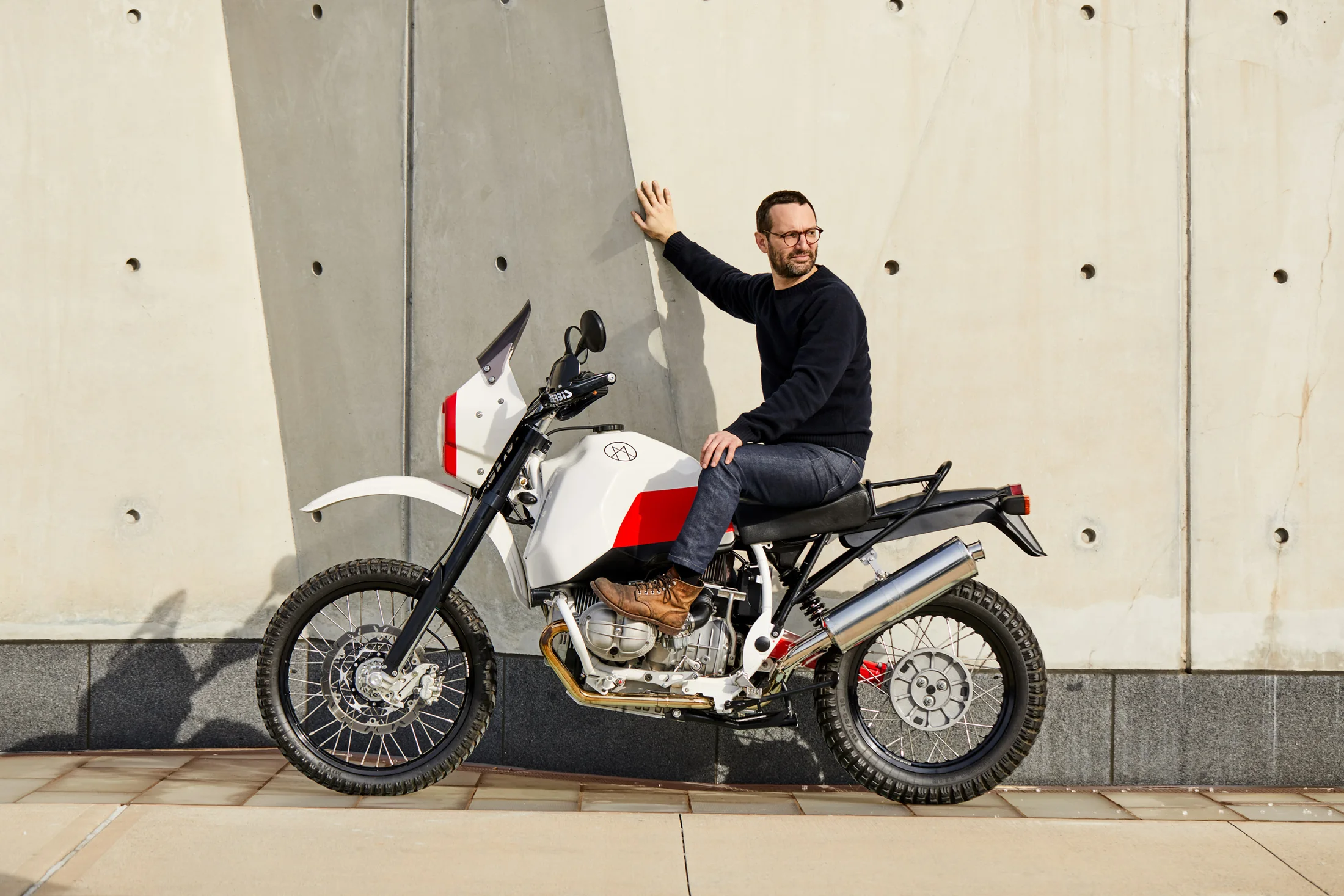 Rider seated on the white and red Myth 001 motorcycle beside a concrete wall