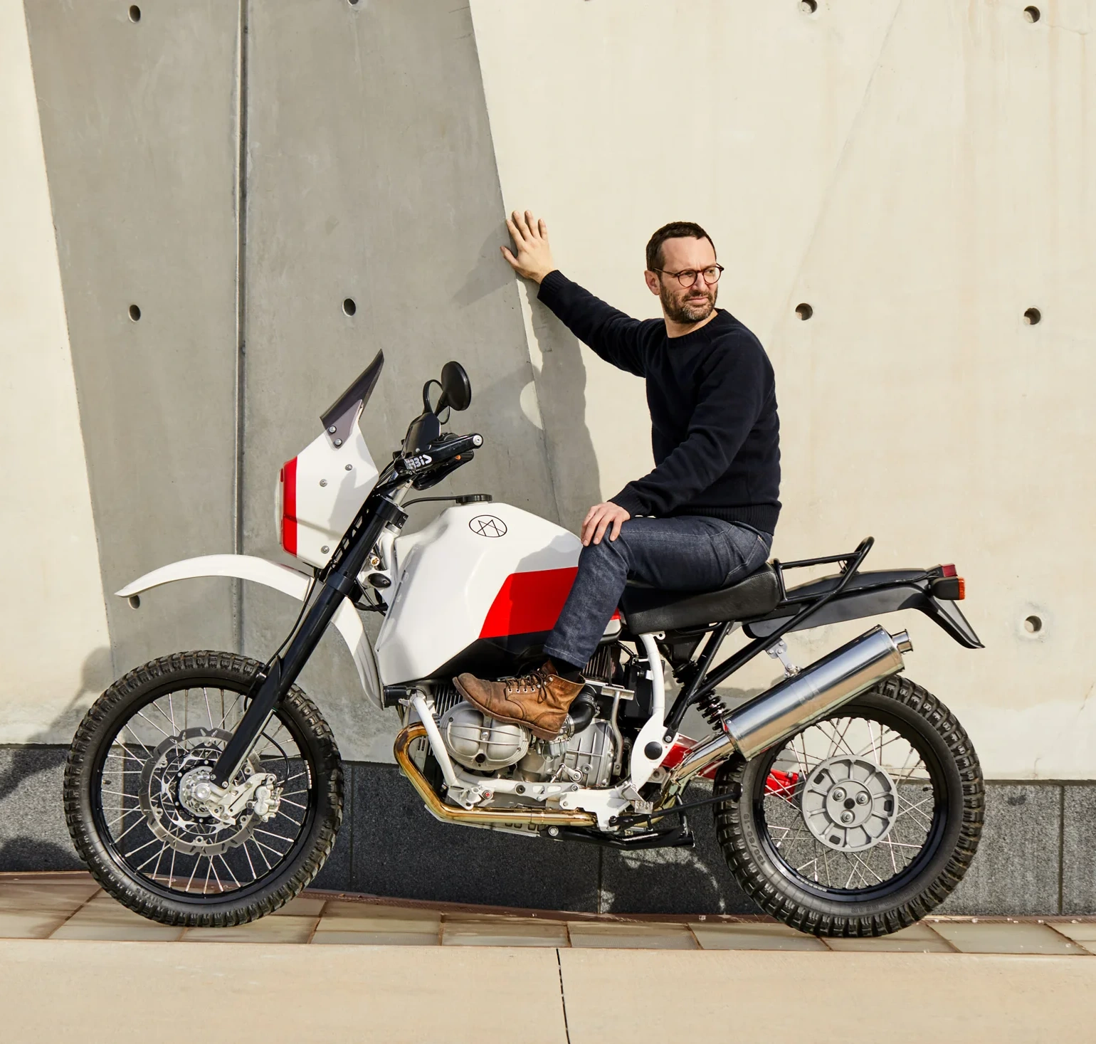 Rider seated on the white and red Myth 001 motorcycle beside a concrete wall