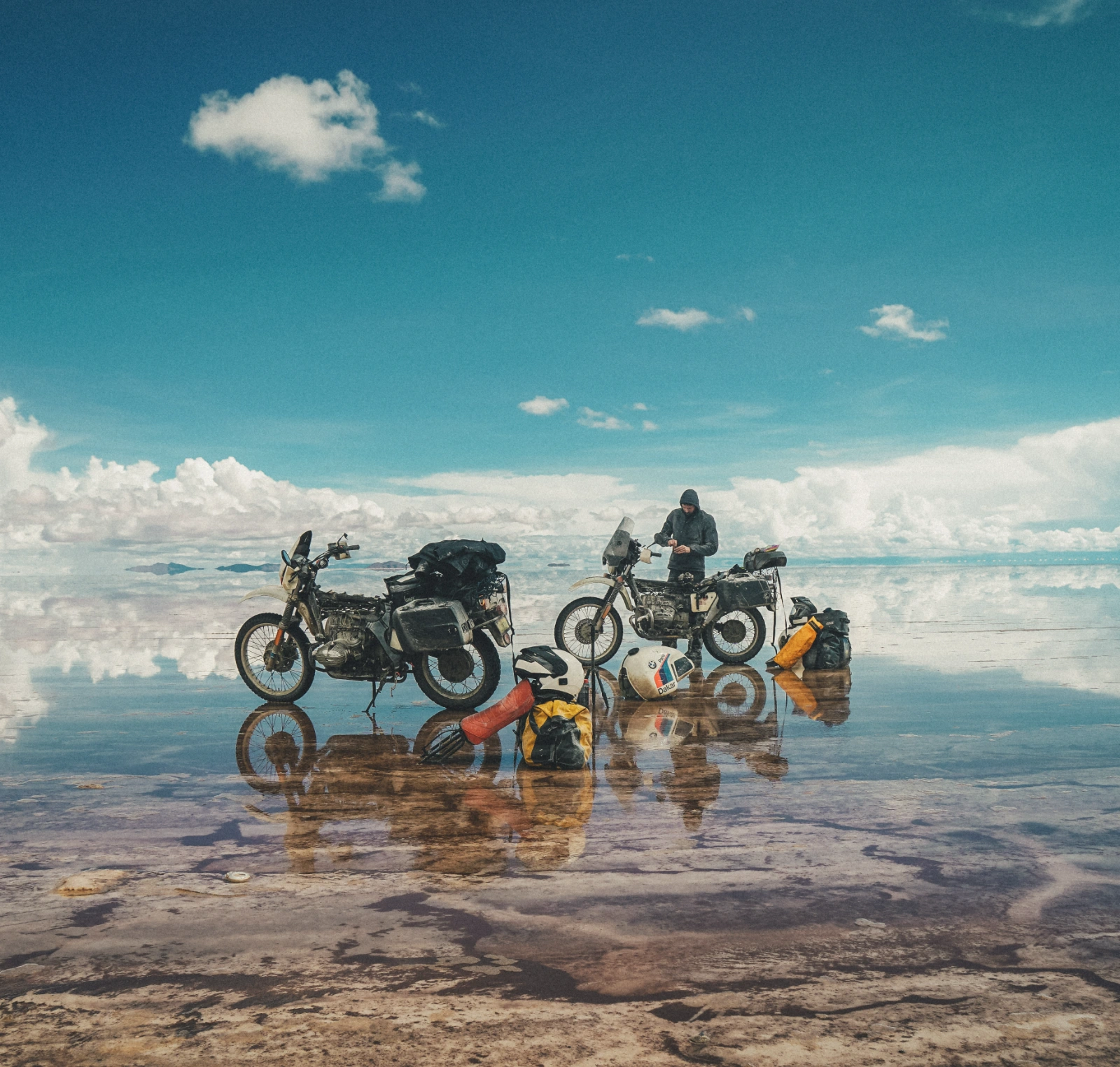 Two loaded motorcycles and travellers reflected in shallow water beneath a blue sky