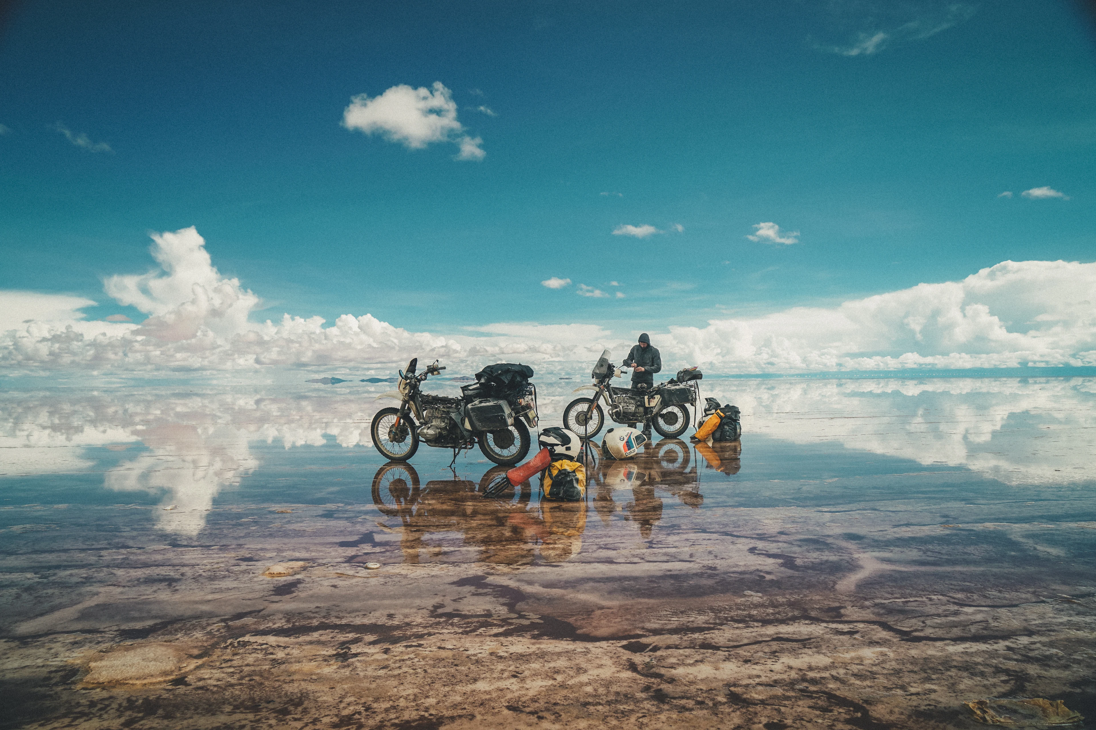 Two loaded motorcycles and travellers reflected in shallow water beneath a blue sky